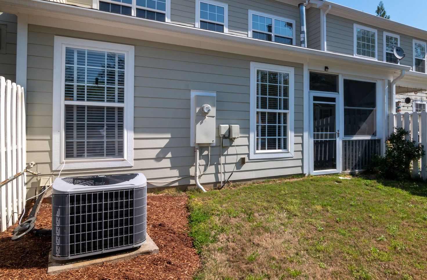 A sunlit backyard view of a modern grey house with white fencing and AC unit.